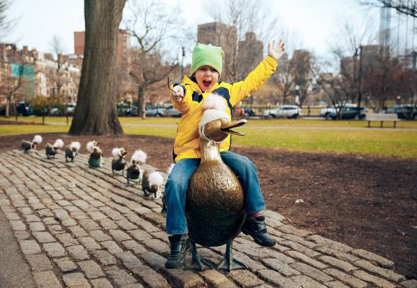 Child sitting on duck statue with line of duckling statues lined up behind it