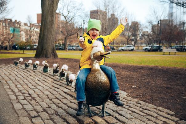 Child sitting on duck statue with line of duckling statues lined up behind it