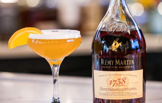 Cocktail glass with orange drink, orange segment, and bottle of cognac on a bar counter