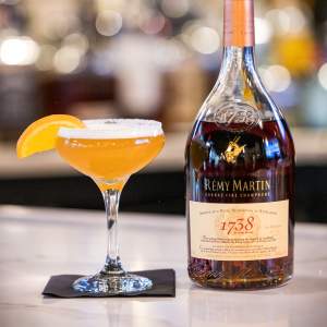 Cocktail glass with orange drink, orange segment, and bottle of cognac on a bar counter