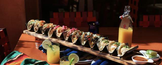Long row of tacos on a board with two citrusy-looking drinks at Rosa Mexicano